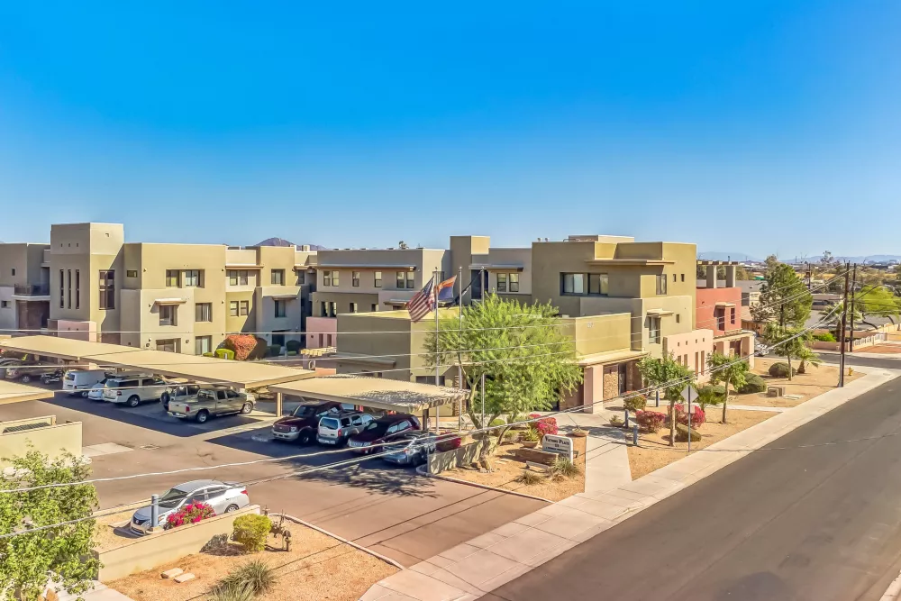Aerial photograph of a desert-style apartment complex with tan buildings, covered parking, and sparse landscaping under a blue sky.