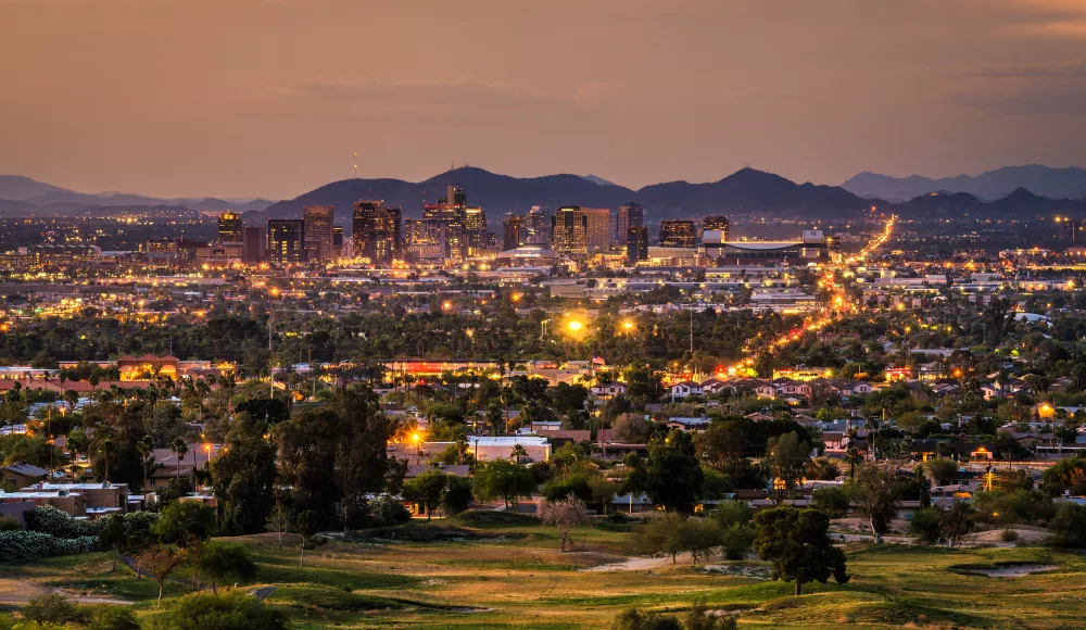 City skyline at dusk, glowing lights illuminate buildings, streets, and homes against distant mountains.