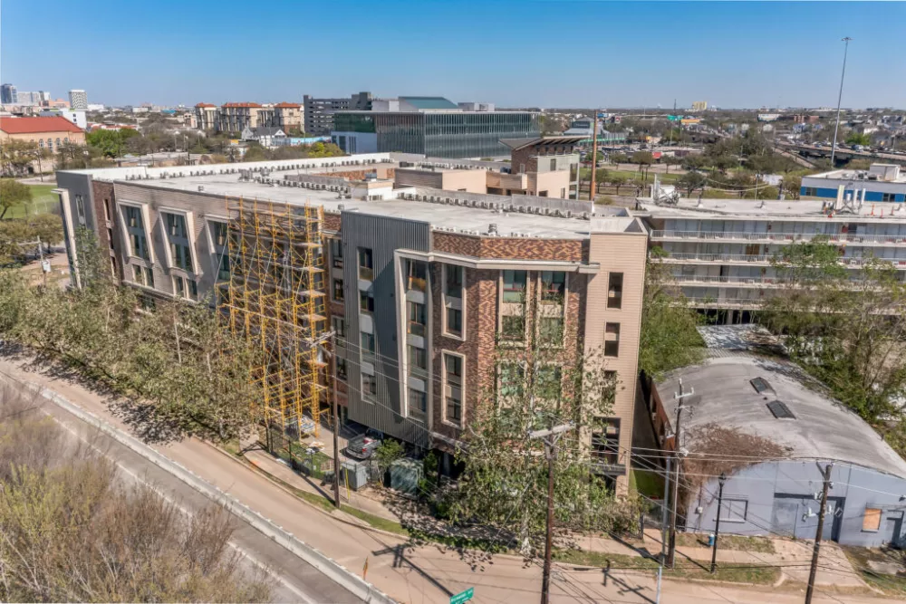 Aerial photograph of a contemporary building with brick, concrete, and yellow lattice details, alongside a street with a city backdrop.