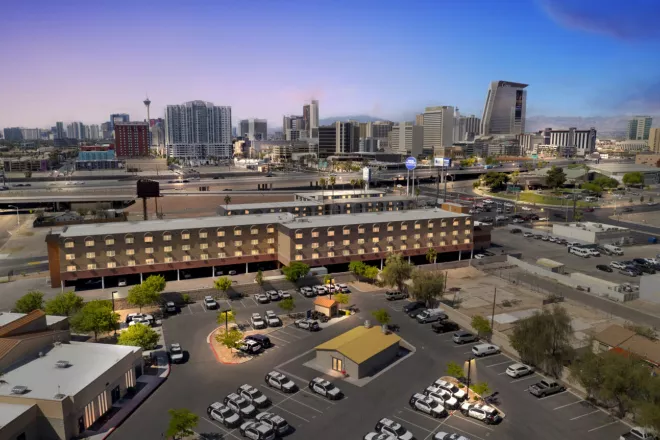 Aerial view of a city, showing a brick hotel with illuminated windows, a full parking lot, a highway, and a distant skyline at dusk.