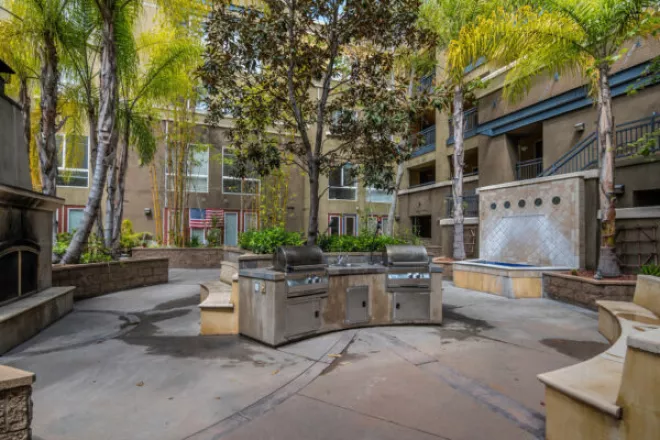 Modern apartment courtyard featuring a double barbecue station, tall palm trees, a waterfall, and outdoor seating.