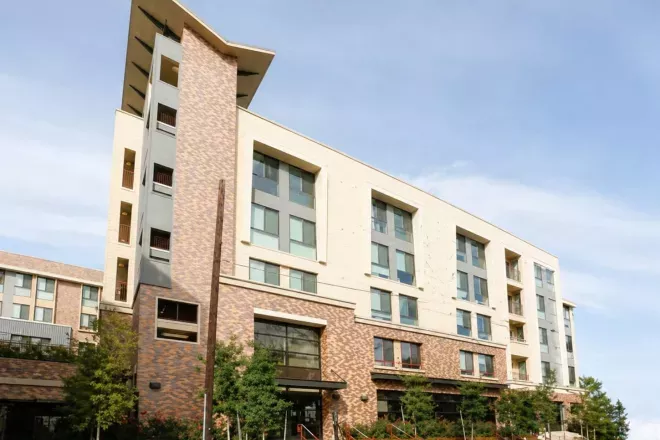 Modern multi-story building with cream, grey, and brick facades, set against a clear sky with some greenery.