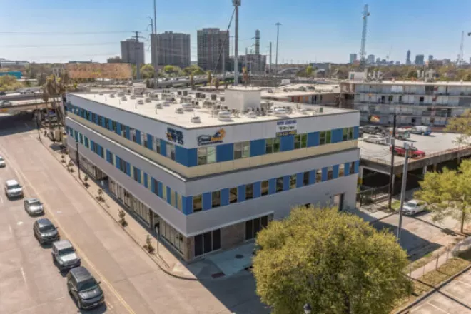 Aerial photograph of a modern three-story building with grey, yellow, and blue accents on a city street corner.