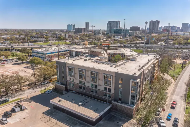 Aerial view of a modern gray apartment building with several cars on a nearby road and a city skyline in the distance.