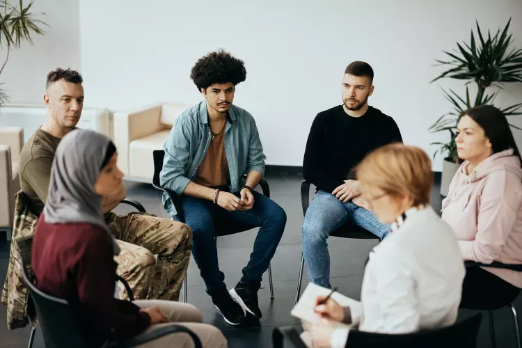 Diverse group of six adults in a circle listening to a facilitator taking notes during a session.